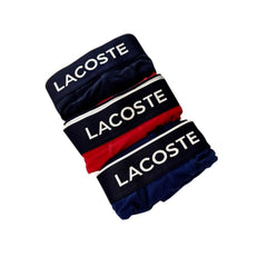 Three pairs of folded Lacoste briefs stacked diagonally on a white background, featuring navy blue, red, and navy blue fabrics with white brand lettering on the waistbands.