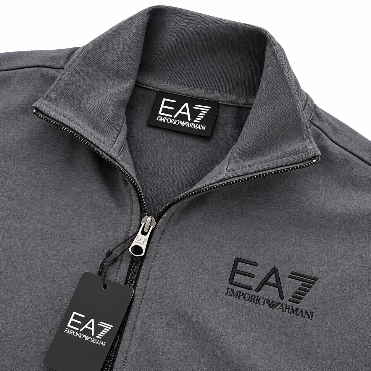 A close-up of a dark gray zip-up jacket by Emporio Armani, featuring the brand's 'EA7' logo embroidered on the left chest and printed on the inner neck label and hanging tag.