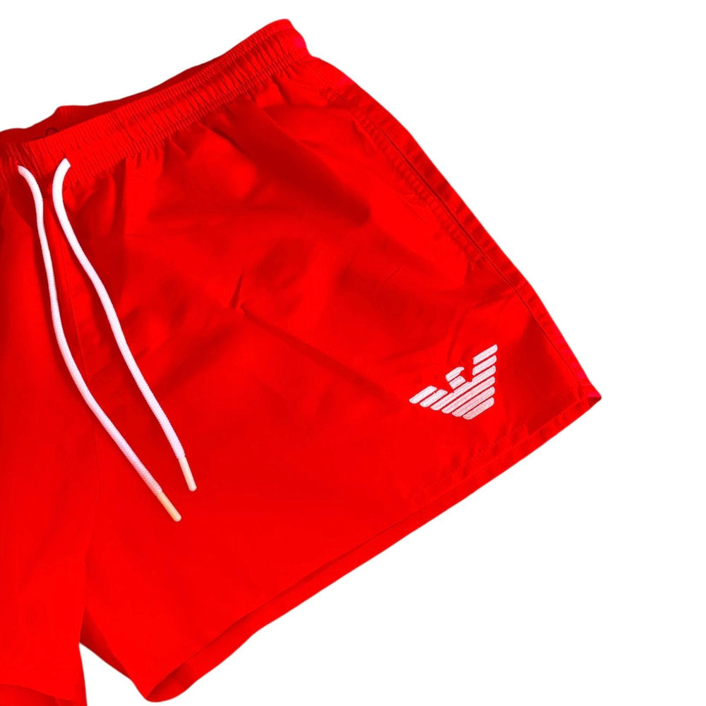Red swim trunks with white drawstrings and a white embroidered eagle logo on the lower left leg, shown against a plain white background.
