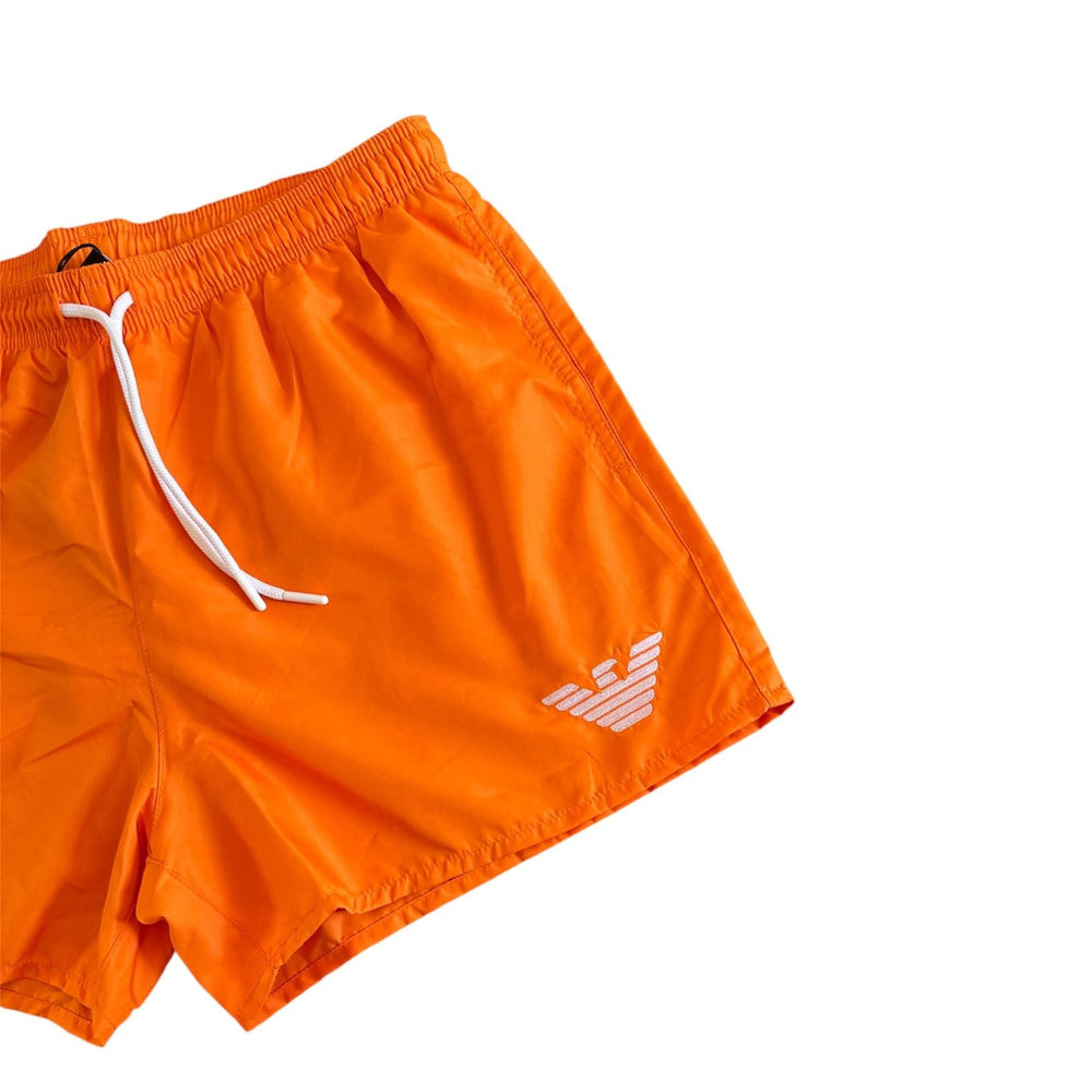 Bright orange swim trunks with a white drawstring and a white embroidered eagle logo on the lower left leg, shown against a plain white background.