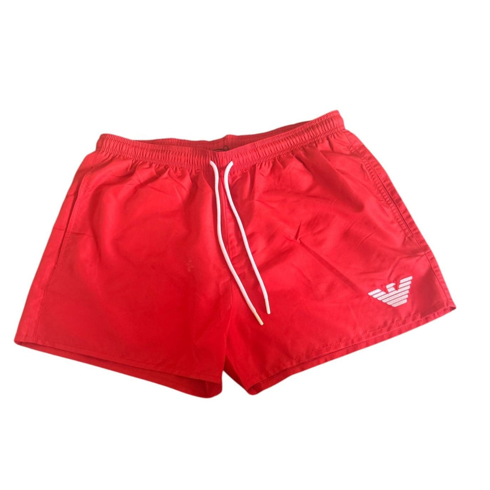 Red swim shorts with a white drawstring and a white embroidered Emporio Armani eagle logo on the lower left leg, shown against a plain white background.